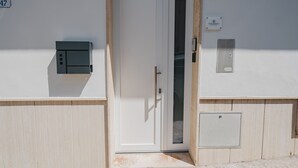 Una puerta blanca con un panel de vidrio, un pequeño cuadro eléctrico y una rejilla metálica en una pared de color claro.