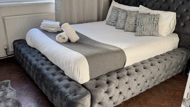 Een slaapkamer met een groot bed, een bekleden hoofdeinde in grijs, witte beddengoed en een grijze plaid.