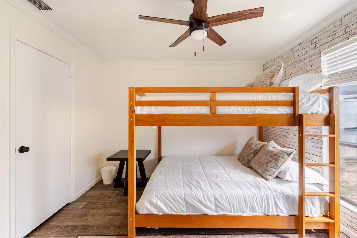 Una cama litera con estructura de madera y ropa de cama blanca, un ventilador de techo, una mesita pequeña y una ventana con persianas.
