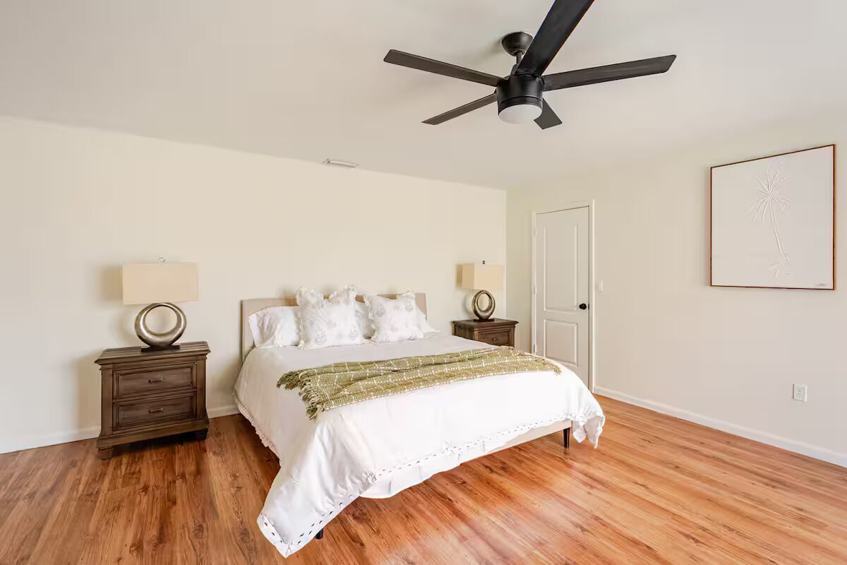 A bedroom with a bed, two bedside tables, a ceiling fan, a door, and a framed picture on the wall.