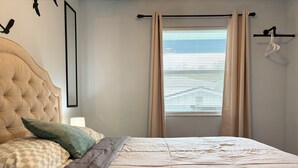 A bedroom with a tufted headboard, a bed with a quilted cover, two pillows, a nightstand with a lamp, a window with blinds, and a coat rack with a hanger.