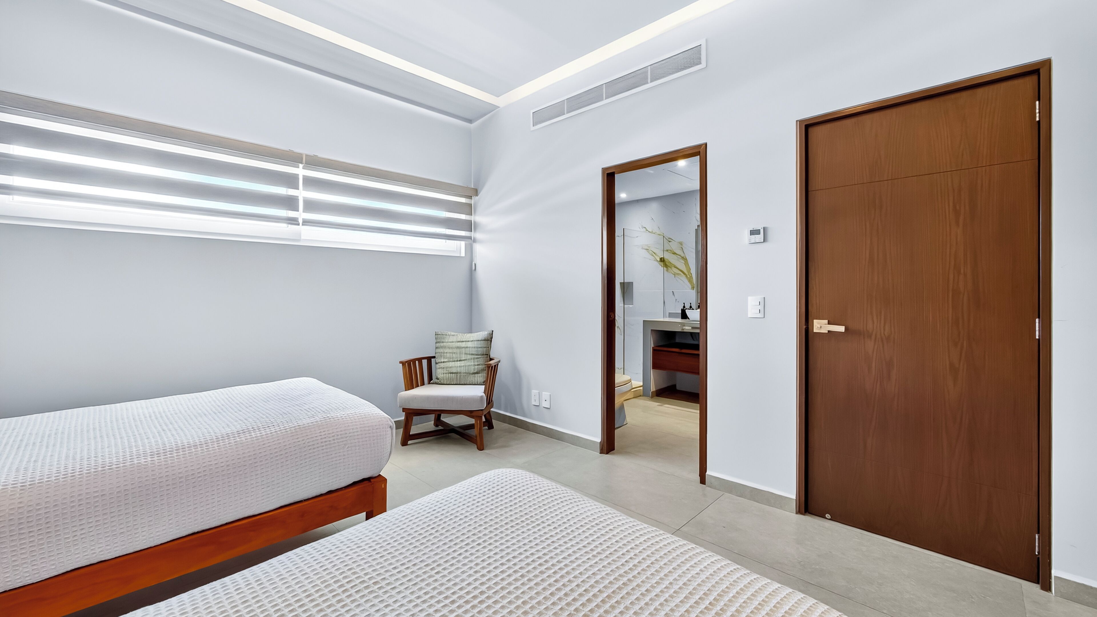 Une chambre avec deux lits, une chaise, une porte en bois et une salle de bain visible à travers une porte ouverte.