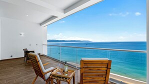 A balcony with a view of the ocean, wooden chairs, a table with glasses and a bottle, and a railing. - Beachfront 2BR - Pools & Balcony Views (Puerto Vallarta)