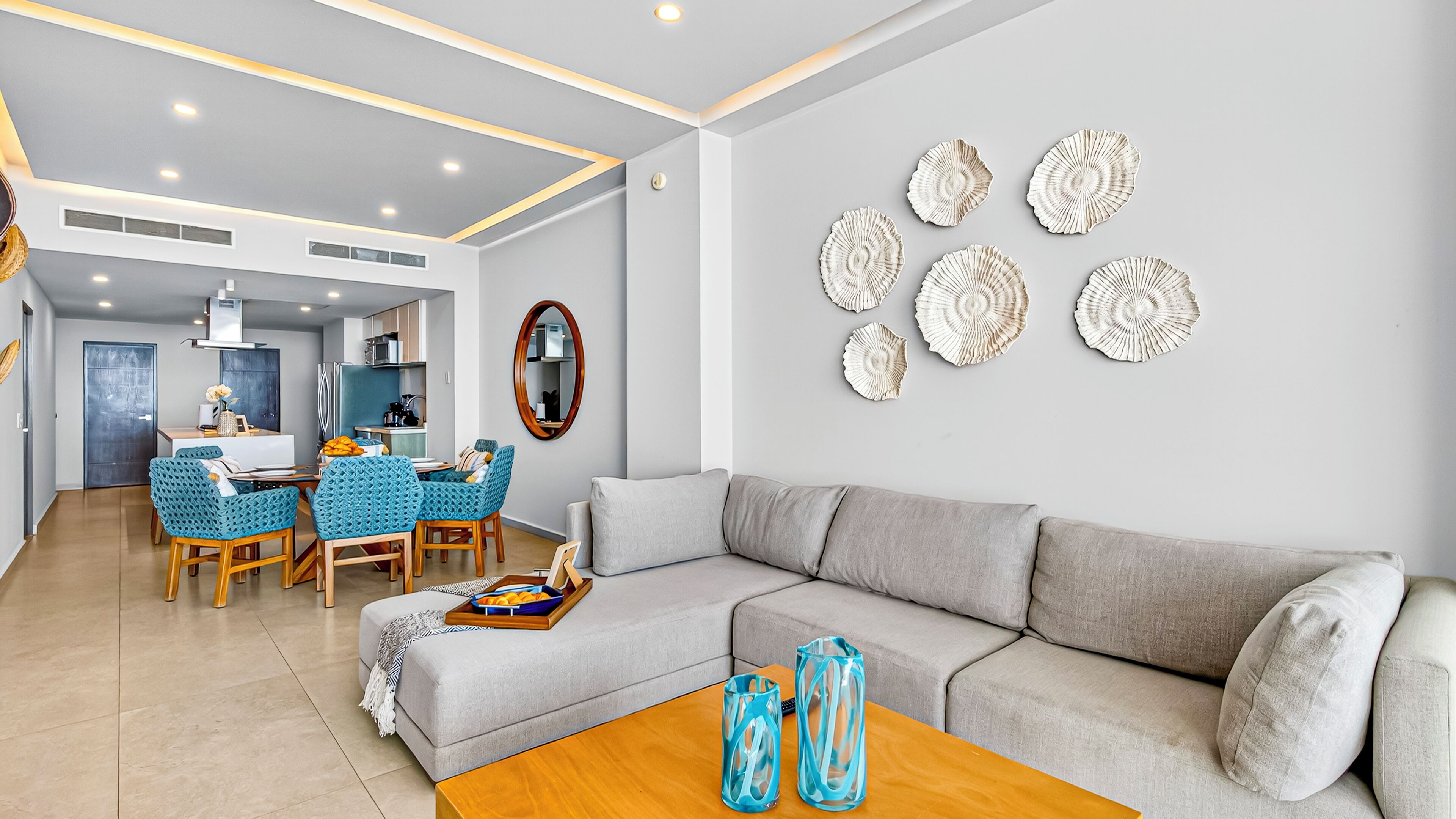 A modern living room with a grey sofa, a wooden coffee table, and a dining area with blue chairs and a kitchen in the background.