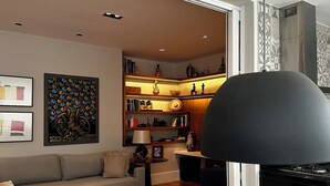 A modern living room with a sofa, ottoman, and a dining table. There is a fireplace and a bookshelf in the background. - Copacabana 3BR • 1 Block to the Beach — Stylish & Family-Friendly (Rio de Janeiro)