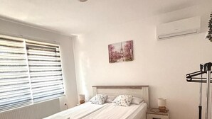 A bedroom with a double bed, a bedside table with a lamp, a radiator, a window with blinds, and a wall-mounted air conditioner. - Alea Place (Sarajevo)
