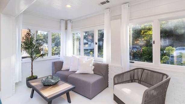 A bright, spacious room with a grey sofa, a wicker chair, and a small table. Large windows offer views of the outside. - Elegant 4BR Oasis in Atlanta! (Atlanta)