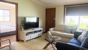A living room with a flat-screen TV on a white stand, a glass coffee table, a blue sofa, a white sofa, and a bed with white bedding.