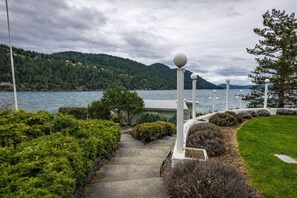 A pathway leading to a building with a green roof, surrounded by lush greenery and a view of a lake with mountains in the background.