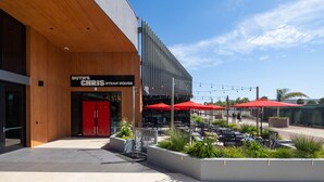 Area salotto esterna con ombrelloni rossi, un ristorante chiamato "Ruth's Chris Steak House" e una moderna facciata di un edificio.