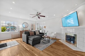 Een moderne woonkamer met een grijze hoekbank, een witte fauteuil, een open haard en een televisie waarop 'Samsung Workspace' wordt weergegeven.