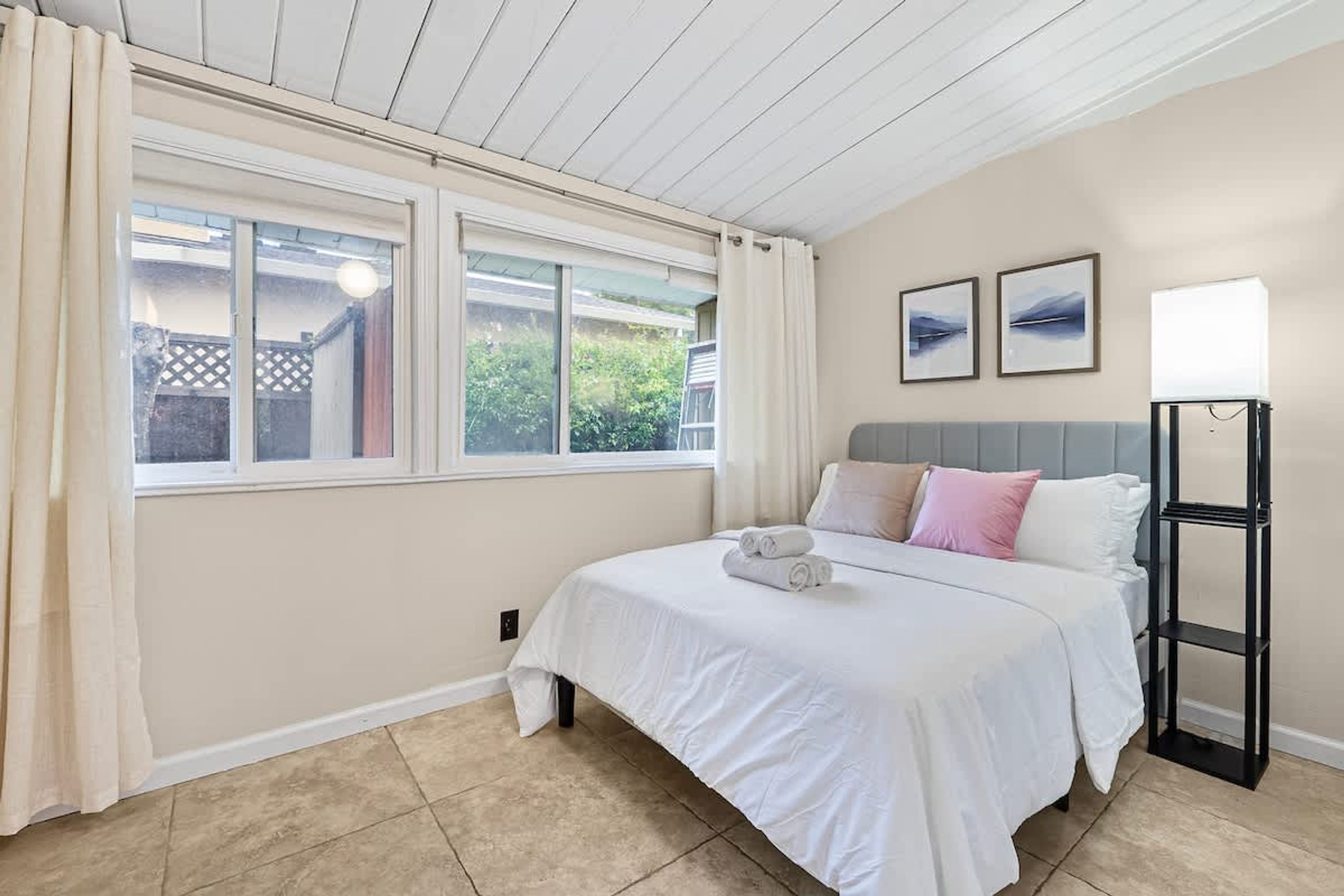 A bedroom with a bed, curtains, a bedside table, a lamp, and two framed pictures on the wall.