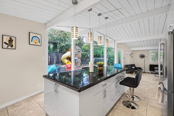 Une cuisine moderne dotée d’un grand îlot central, de luminaires suspendus et offrant une vue sur un jardin avec un espace de jeux pour enfants.