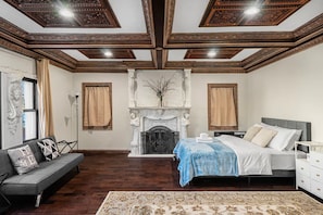 A bedroom with a fireplace, a bed with a blue bedspread, a sofa with cushions, and a patterned rug.