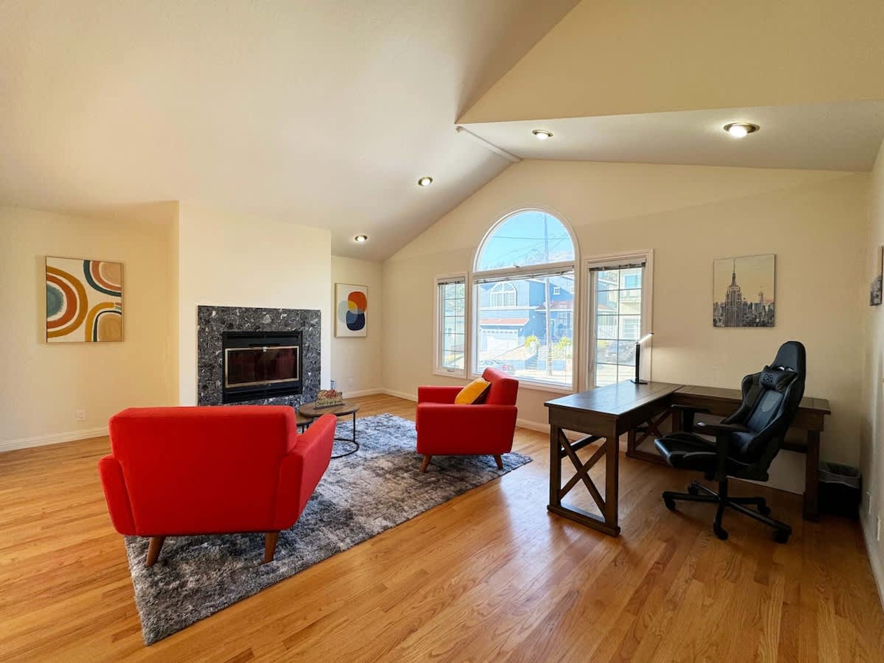 A spacious living room with a fireplace, red chairs, a desk, and large windows.