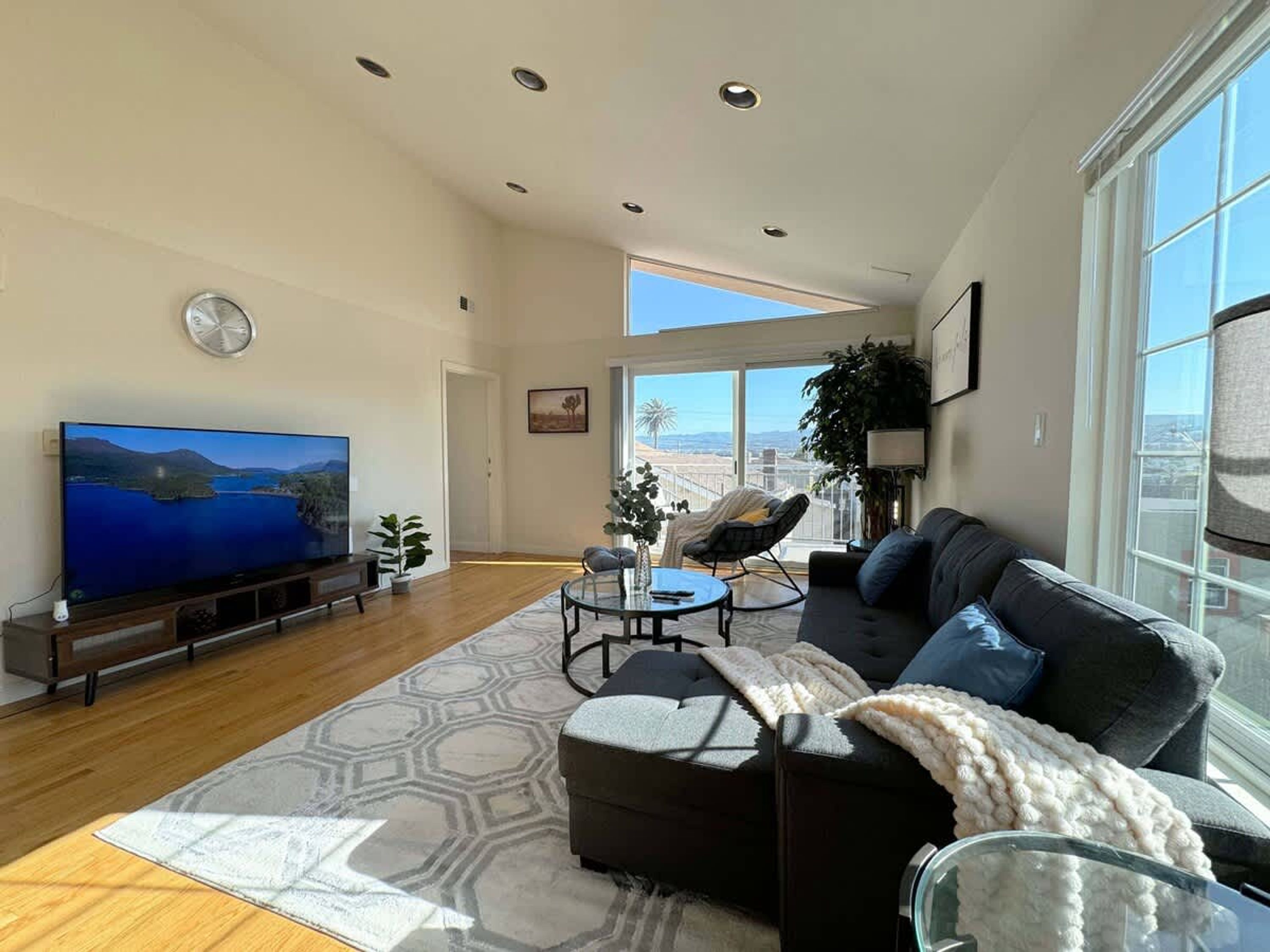 A spacious living room with a large flat-screen TV, a dark-colored sofa, a glass coffee table, and a patterned rug.