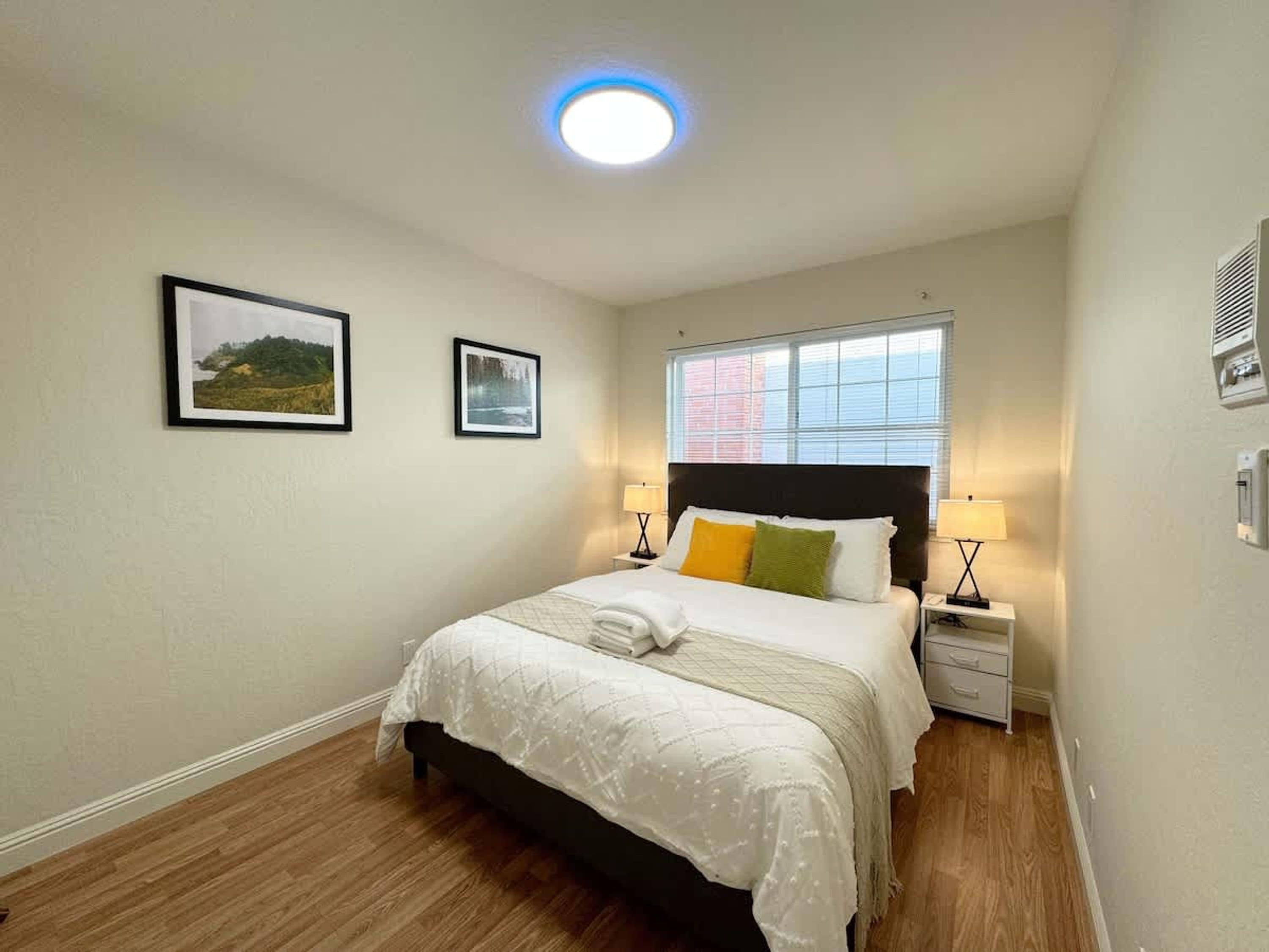 A bedroom with a bed, two bedside tables, a window, and two framed pictures on the wall.
