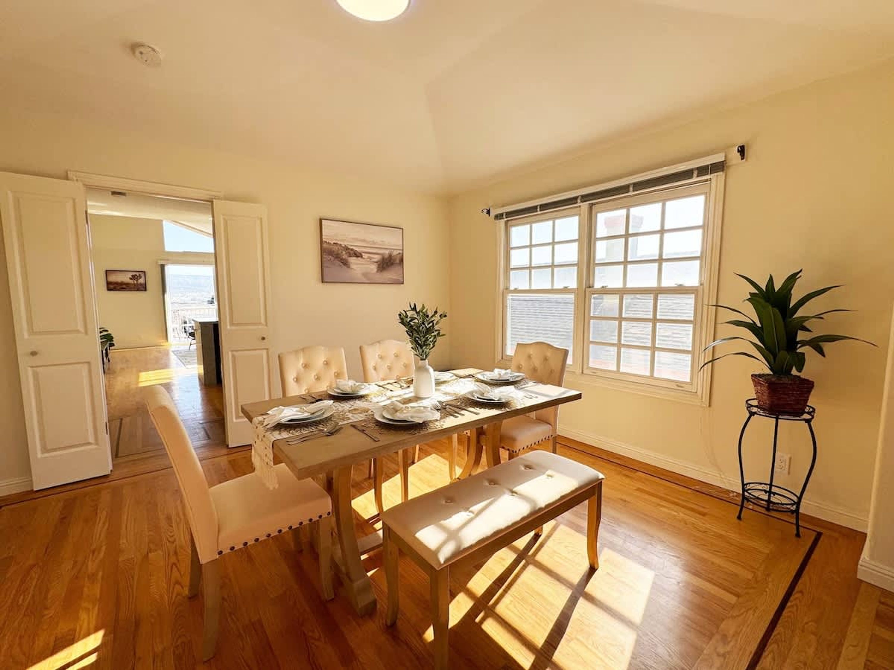 A dining area with a wooden table set for four, surrounded by cushioned chairs. There is a bench in the center, a potted plant on a stand, and a painting on the wall.