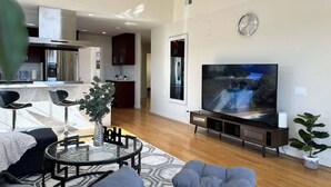 Ein modernes Wohnzimmer mit Flachbildfernseher, Glastisch und einer Sitzecke mit Sofa und Hocker.