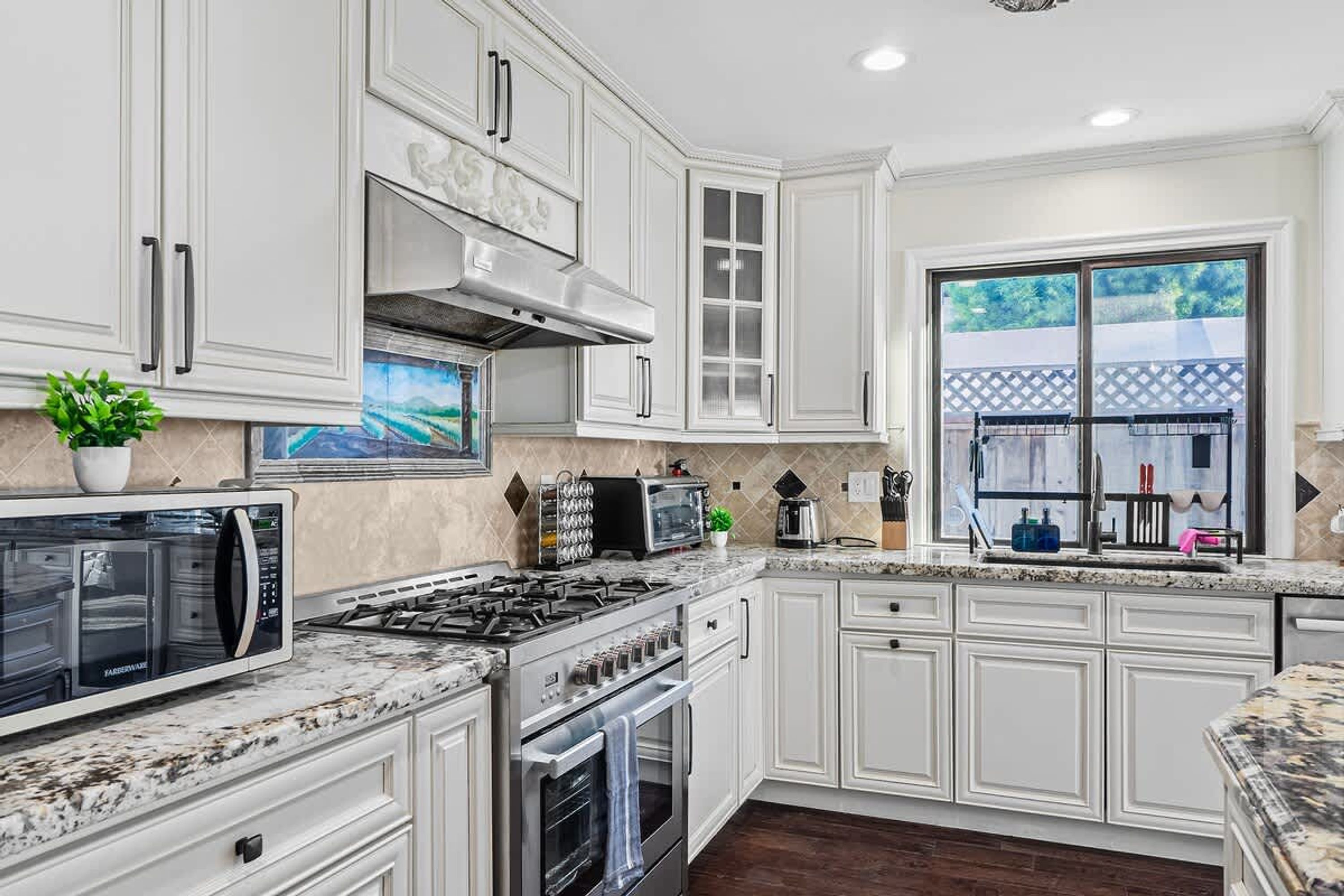A modern kitchen with white cabinetry, a gas stove, an oven, a microwave, and a large window offering a view of the outdoors.