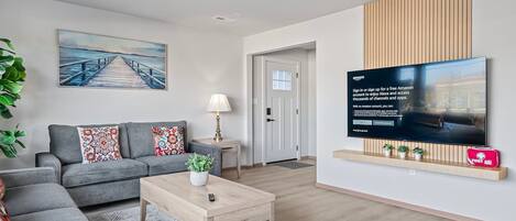 A modern living room with a grey sofa, a coffee table, and a painting of a pier. There's a TV mounted on the wall displaying an Amazon sign.