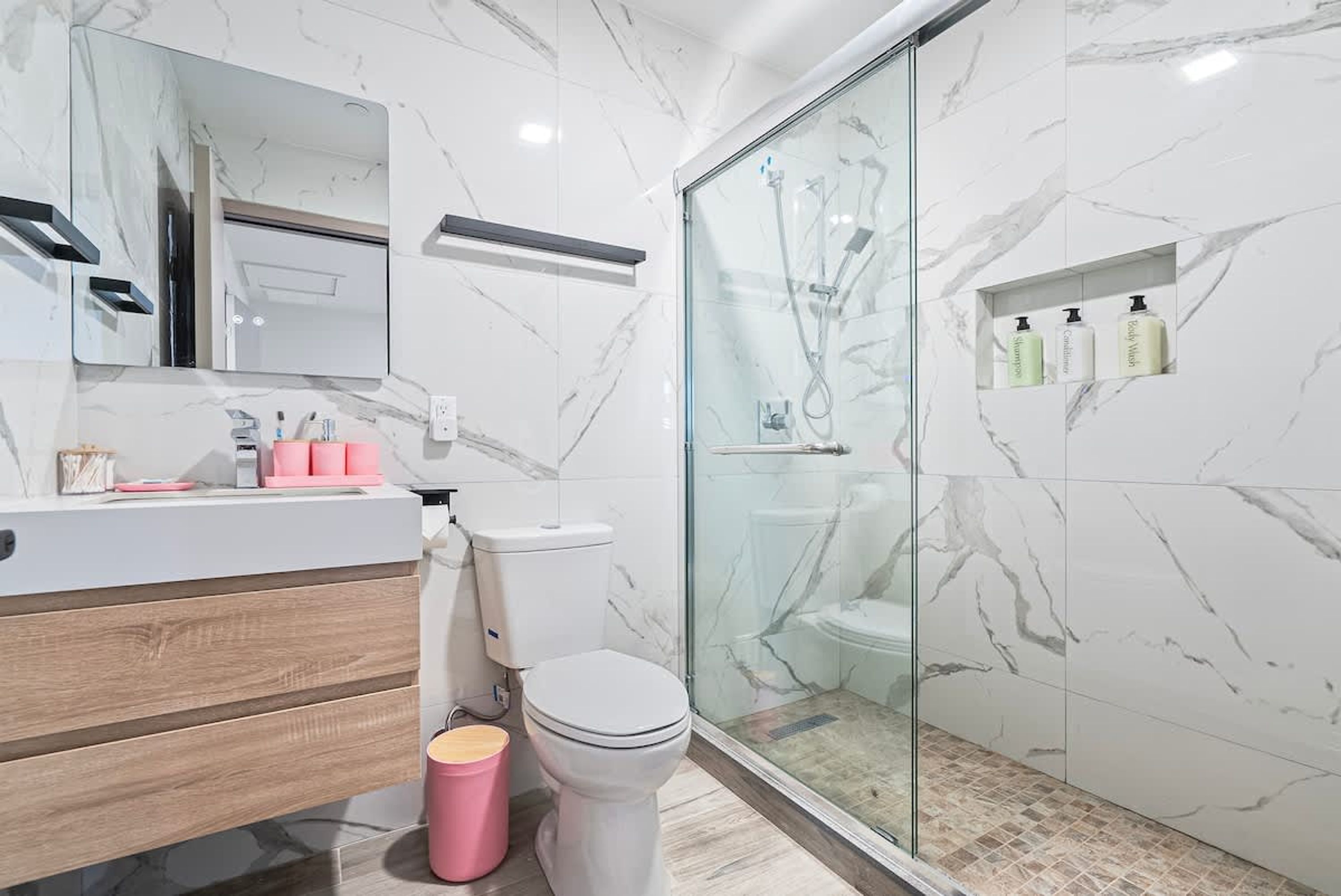 A modern bathroom with a marble backsplash, a glass shower enclosure, and a vanity with a mirror and toiletries.
