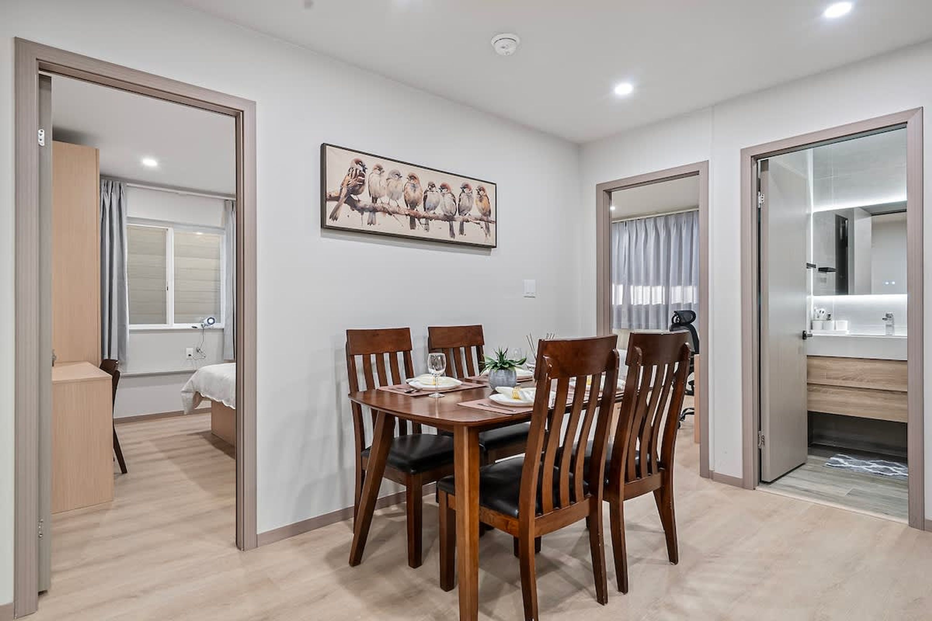 A dining area with a wooden table and chairs, a painting of birds on the wall, and a kitchen area visible through an open door.