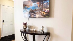 A room with a wall-mounted TV displaying a scenic image, a small round table with a decorative pig, a lamp, and a picture frame. - Campbell Cozy Bright 2B 1b AC Close to Downtown (Campbell)