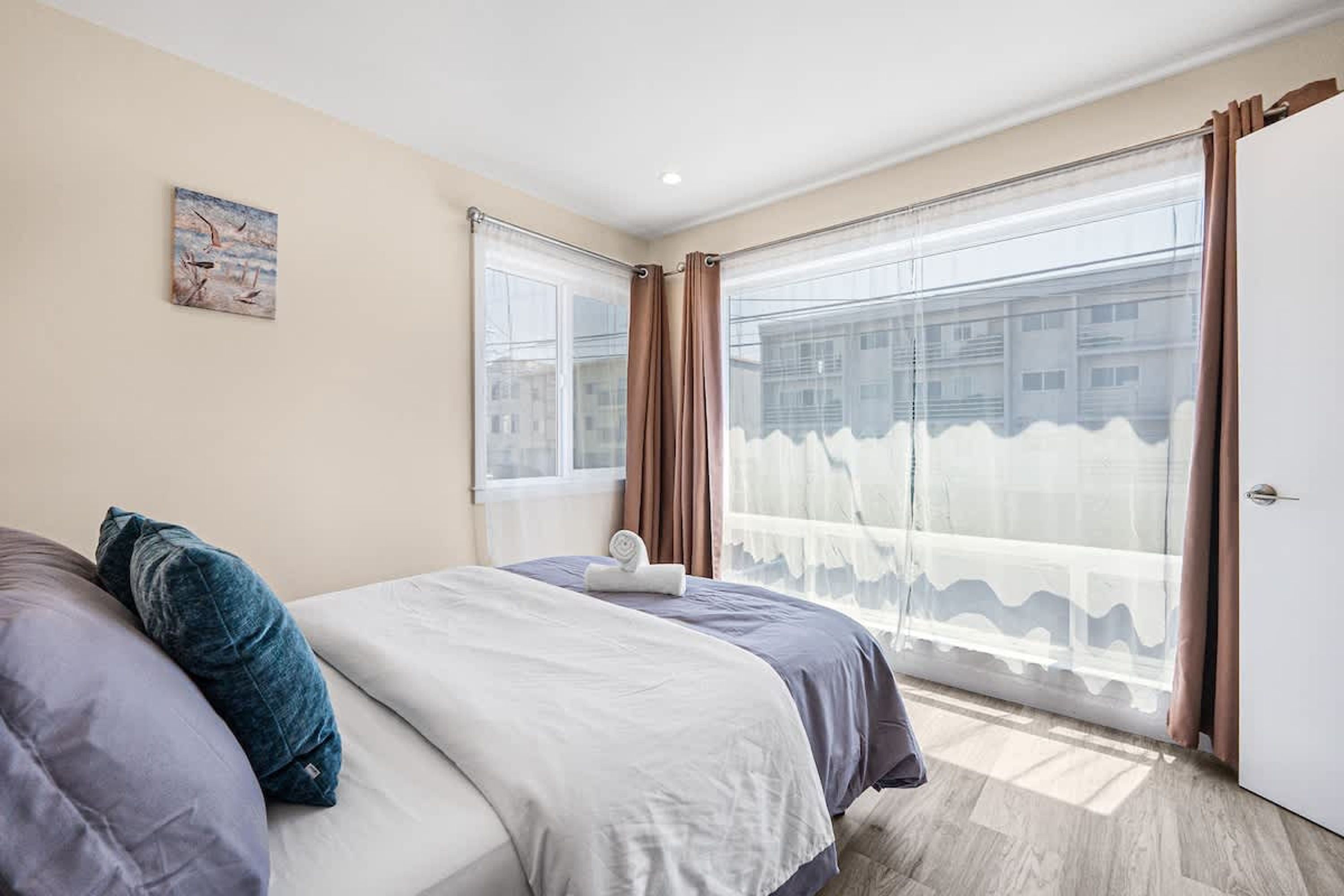 A bedroom with a bed, a window with curtains, a painting on the wall, and a door.