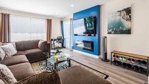 Ein modernes Wohnzimmer mit einer Eckcouch, einem Glastisch, einem Kamin, einem Fernseher an einer blauen Akzentwand und einem Schuhregal.