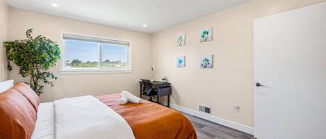 Een slaapkamer met een bed, bureau, stoel, een kamerplant en kunst aan de muur.
