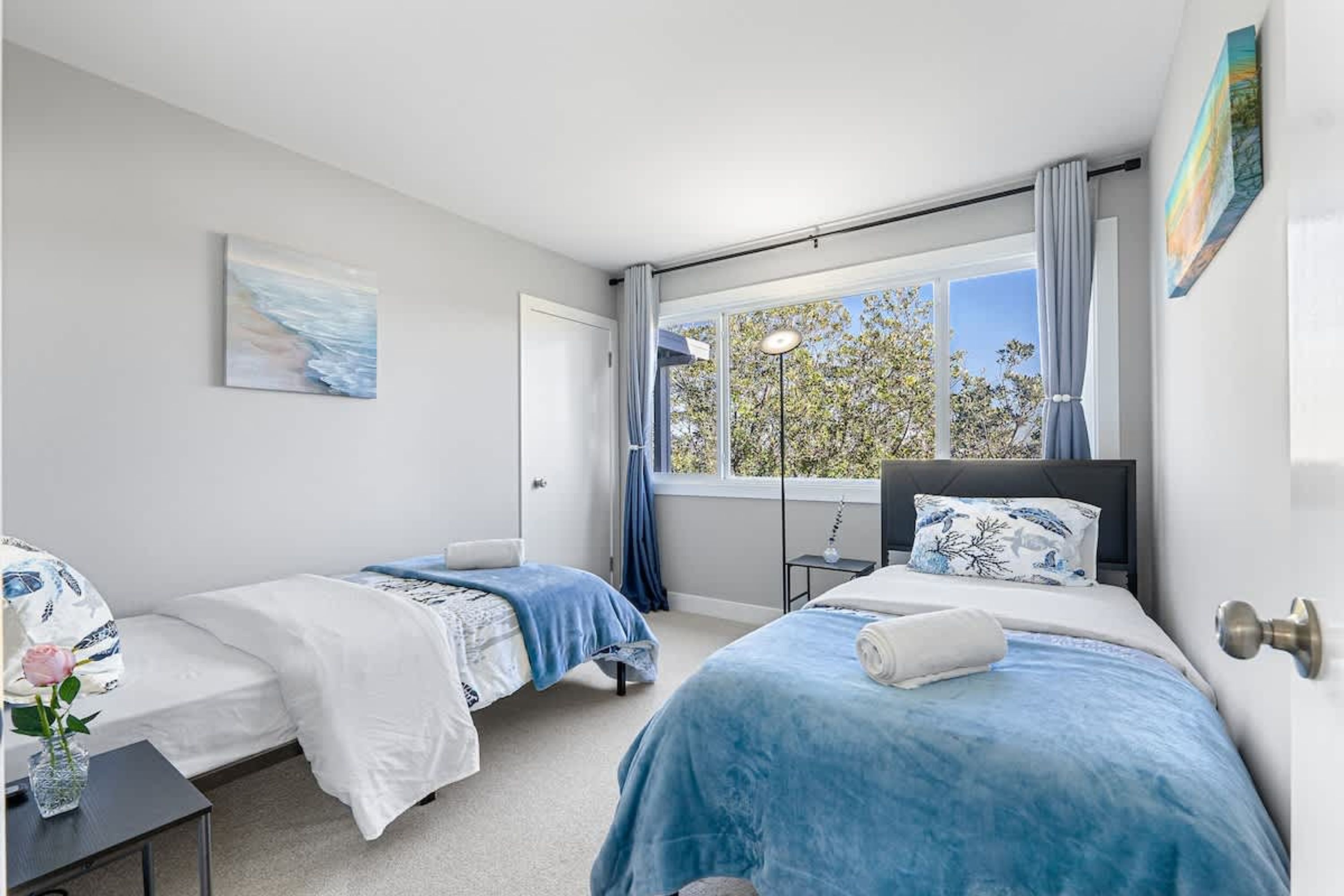 A hotel room with two beds, a large window with curtains, a bedside table with a lamp, and a painting on the wall.