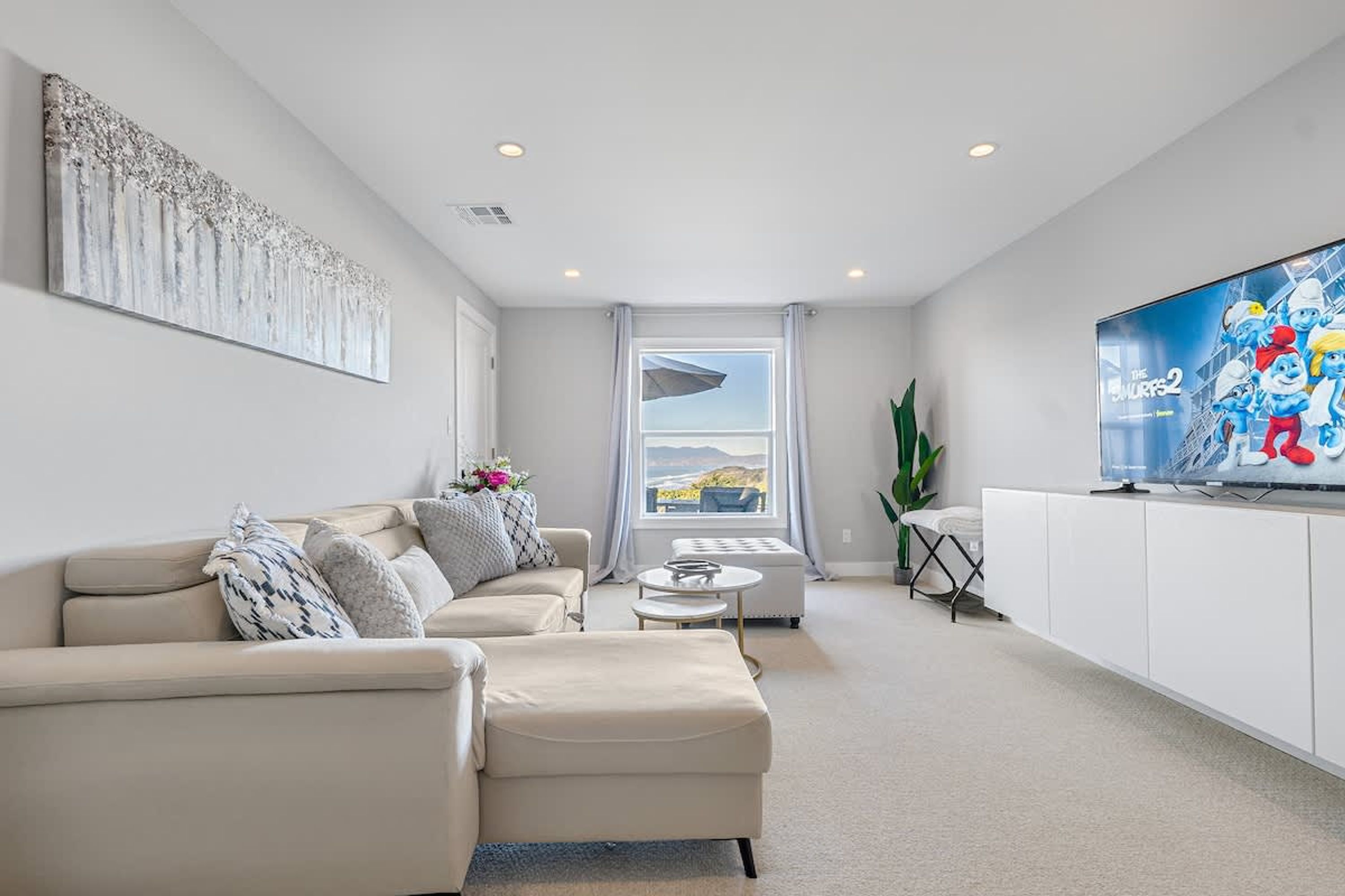A modern living room with a beige sofa, a coffee table, and a flat-screen TV displaying 'The Smurfs 2'. A large window offers a view of the outdoors.
