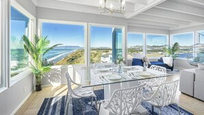 Un comedor amplio con una mesa de vidrio y sillas blancas, ventanas grandes que ofrecen vistas al océano y las colinas, y una lámpara de araña moderna.
