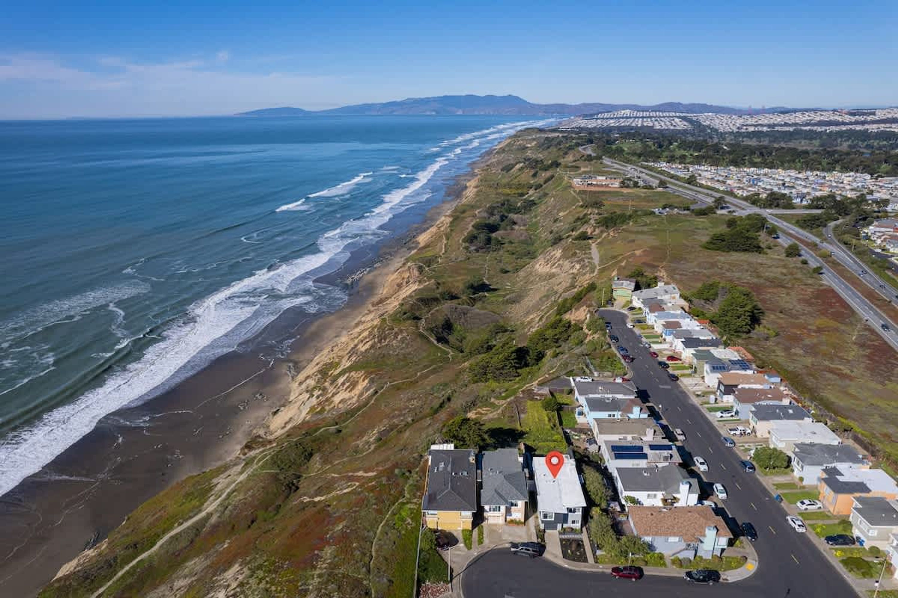 An aerial view of a coastal residential area with houses along the beach, a road, and a hilly landscape in the background.