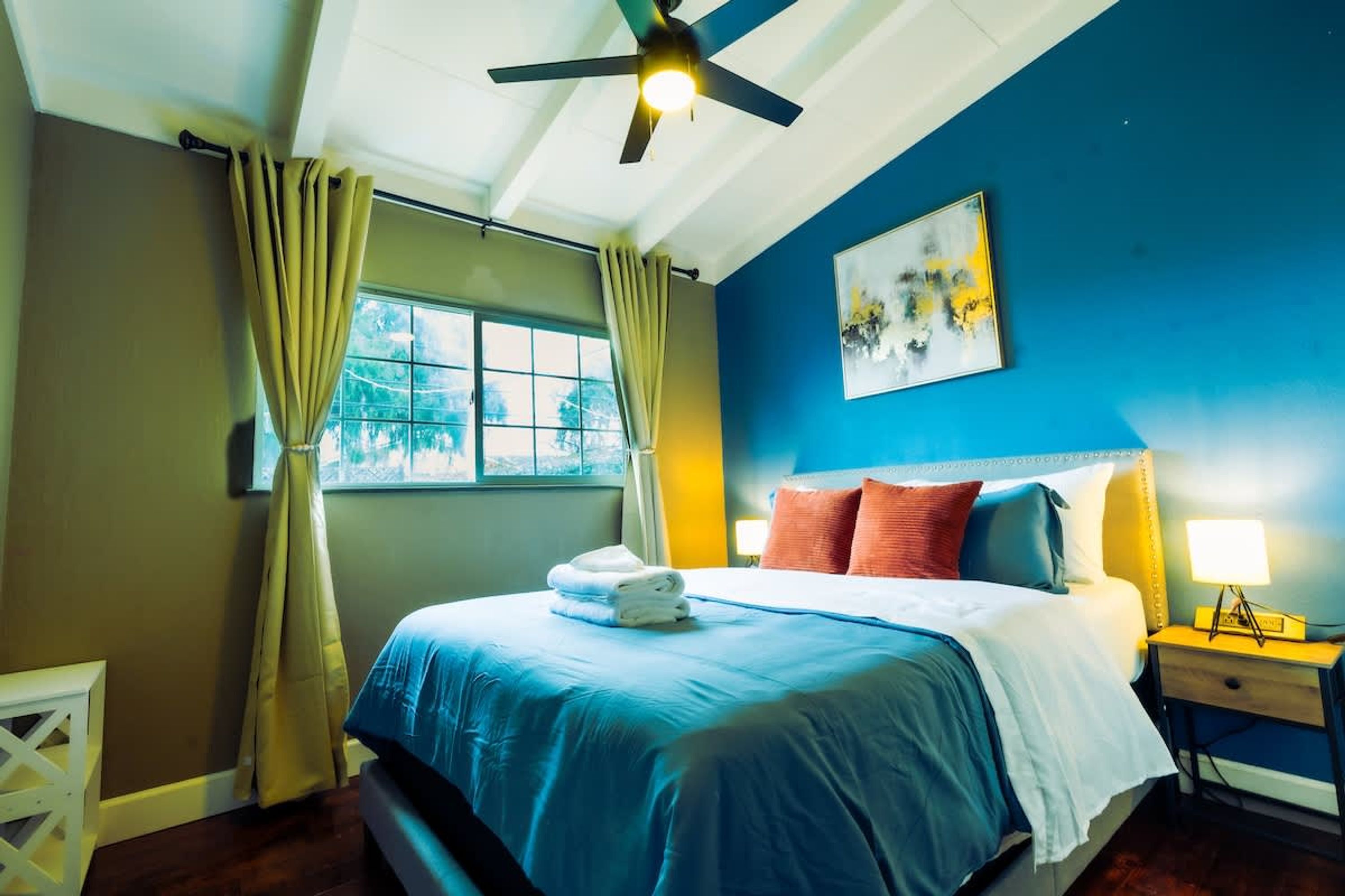 A bedroom with a bed, a ceiling fan, a window with curtains, a bedside table with a lamp, and a painting on the wall.