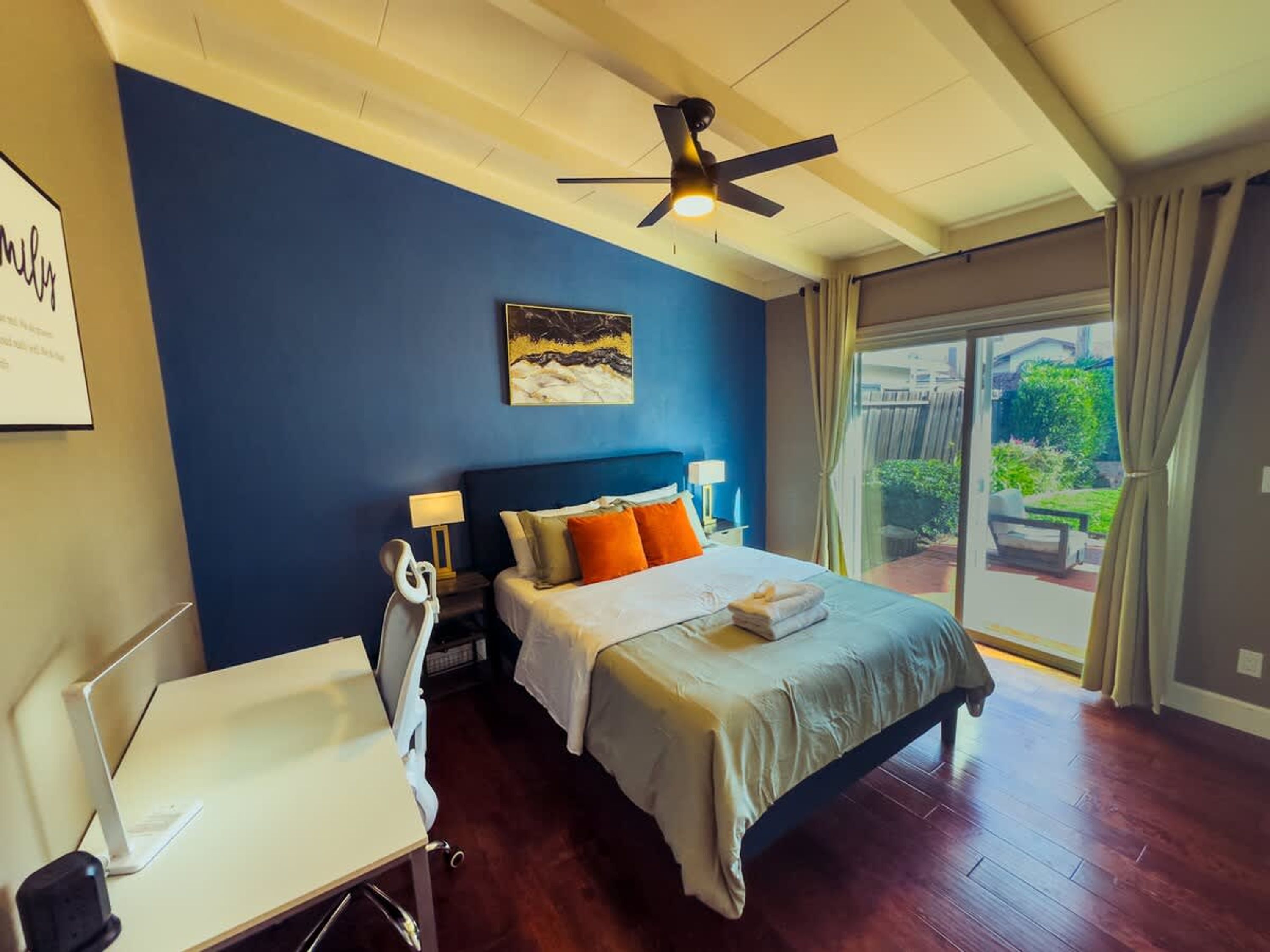Una habitación de hotel con cama, escritorio, silla, ventilador de techo y un amplio ventanal con cortinas.