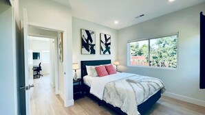 A bedroom with a bed, a nightstand, two wall-mounted abstract paintings, a window with a view of greenery, and a door leading to another room. - New 3b3b Home Gogle Speed Wifi (Sunnyvale)