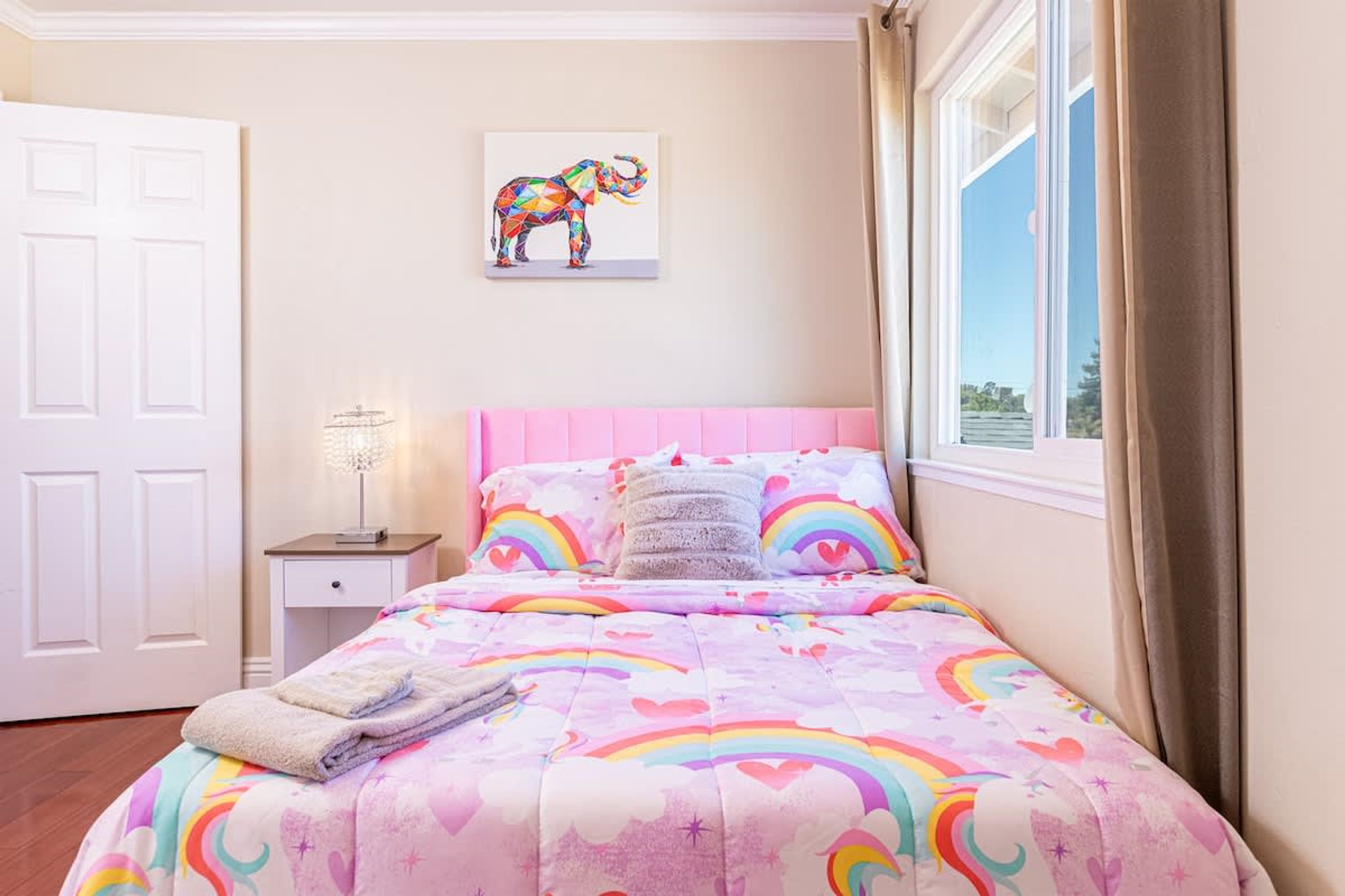 Un dormitorio con una colcha rosa adornada con motivos de arcoíris y corazones, una puerta blanca, una mesita de noche con una lámpara y una ventana con cortinas.