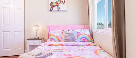 Un dormitorio con una colcha rosa adornada con motivos de arcoíris y corazones, una puerta blanca, una mesita de noche con una lámpara y una ventana con cortinas.