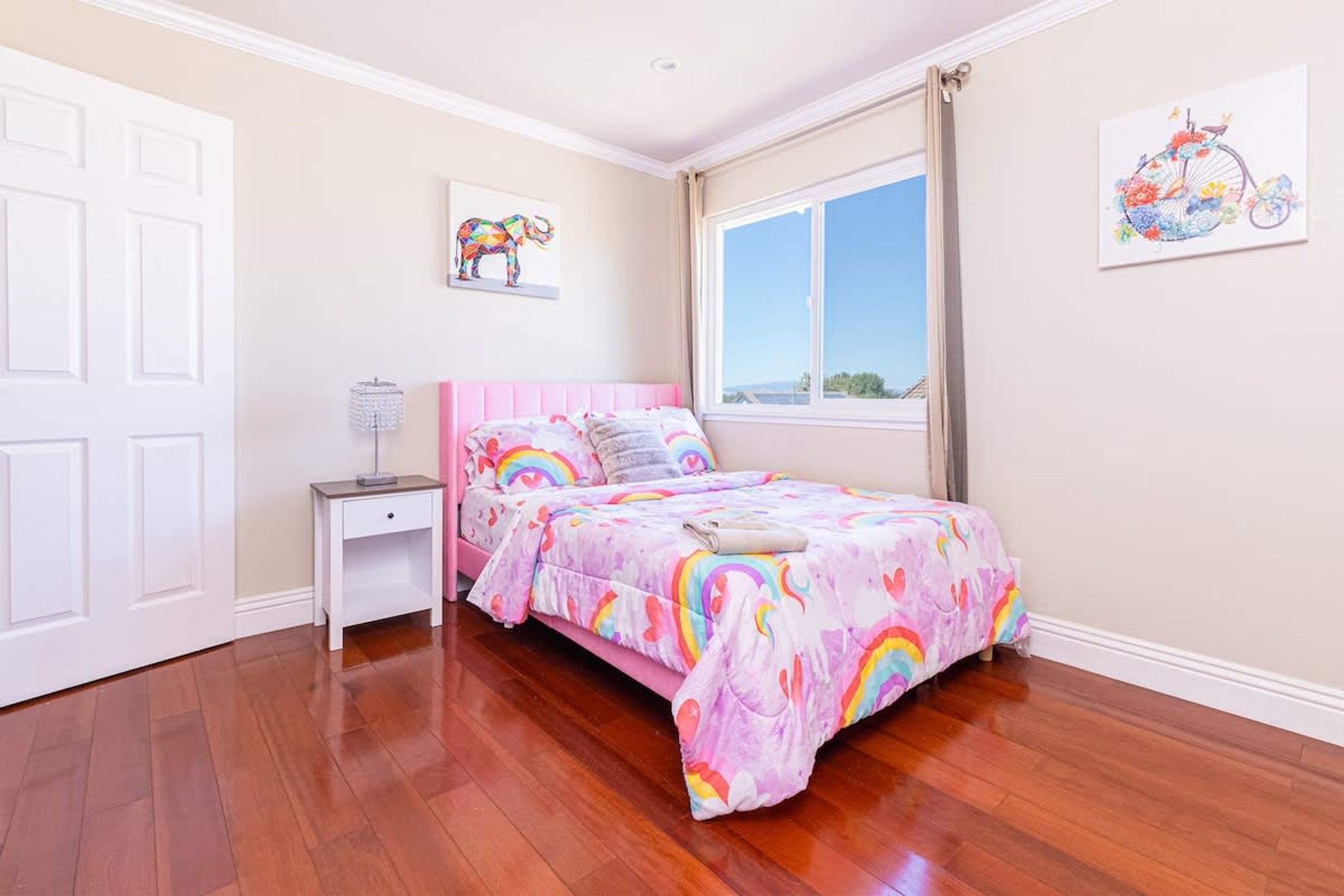 A bedroom with a pink bed, a nightstand, a window with a view, and a door.
