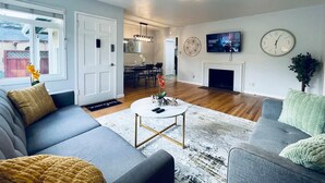 Un salon moderne avec un canapé gris, une table basse et une cheminée. On aperçoit à l’arrière-plan un coin repas agrémenté d’un lustre.