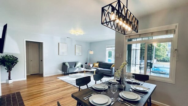 Un espace de repas moderne avec une table en bois dressée pour quatre personnes, un canapé gris en arrière-plan et une grande fenêtre donnant sur l’extérieur.