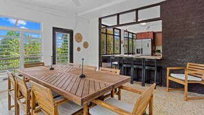 A spacious dining area with a wooden table and chairs, a kitchen with a brick wall, and large windows offering a view of greenery.