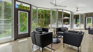 A modern living room with black leather sofas, a round coffee table, and a ceiling fan. Large glass doors with metal shutters lead to a garden area.