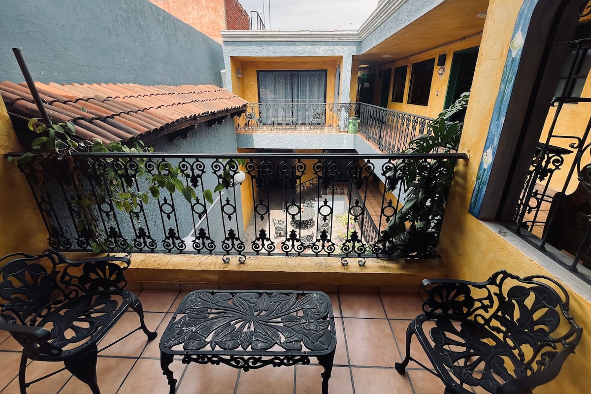 A balcony with a tiled floor, two black wrought iron chairs, a small table, and a railing with plants.