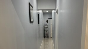 A long, narrow hallway with light grey walls and a shiny tiled floor. There is a metal rack with shelves against the wall, and a door at the end of the hallway. - Cozy air conditioned 2-bedroom apartment in charming Charles Town community. (Charles town)