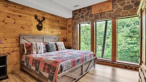A wooden-paneled bedroom with a large bed, stone wall, and a window overlooking a forest. - Mountain Views, 🎯Game Room, Sleeps 20, Near Dollywood & Gatlinburg (Sevierville)