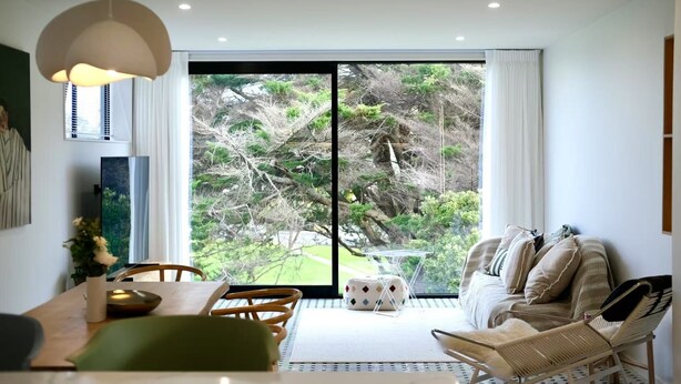 Een moderne woonkamer met een grote schuifpui naar een tuin, een eethoek met een tafel en stoelen, en een gezellige zithoek met een bank en fauteuils.