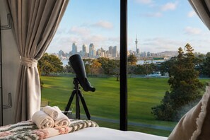 A telescope on a tripod in a room with a bed and curtains, overlooking a cityscape and park. - Stylish House with Great City Views (Auckland)